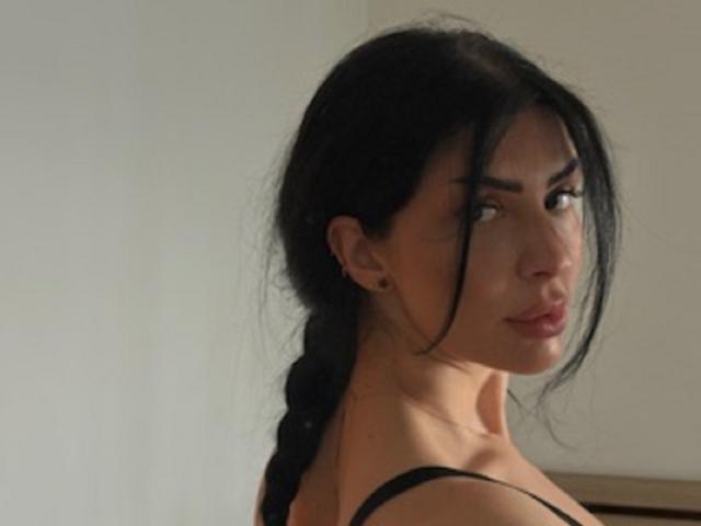 BrainyBelle with black braided hair, looking over shoulder, showing her tits in a black strap top, serious expression, soft light, pale background.