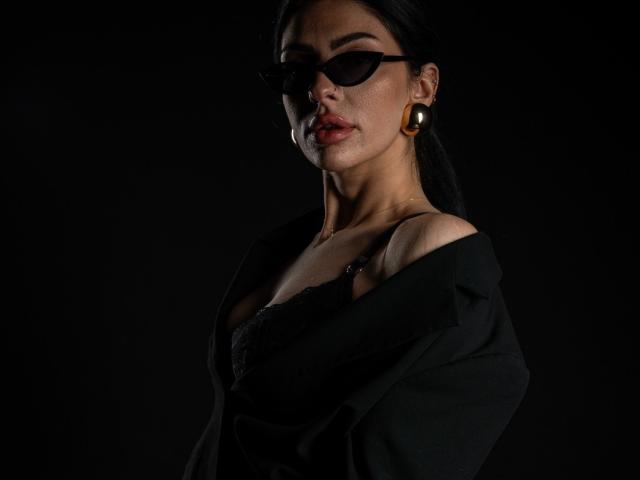 BrainyBelle with dark sunglasses, black lace bra, and black jacket off-shoulder, showing her tits, wearing large gold earrings, dark hair, and a serious expression, against a black background.