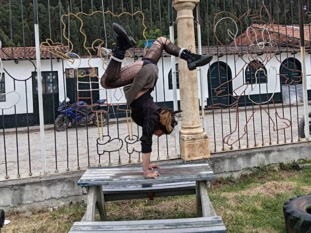 SakuraBlackRose está de cabeça para baixo em um banco, vestindo uma camisa preta, calças marrons e botas pretas, com o traseiro para cima e a cabeça entre as pernas..