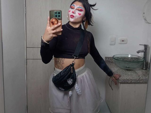 SakuraBlackRose avec un visage peint en blanc, un haut noir à manches longues, une jupe blanche, un sac noir, tenant un téléphone, debout dans une salle de bain, montrant des tatouages, tenant la jupe relevée, poitrine moyenne..