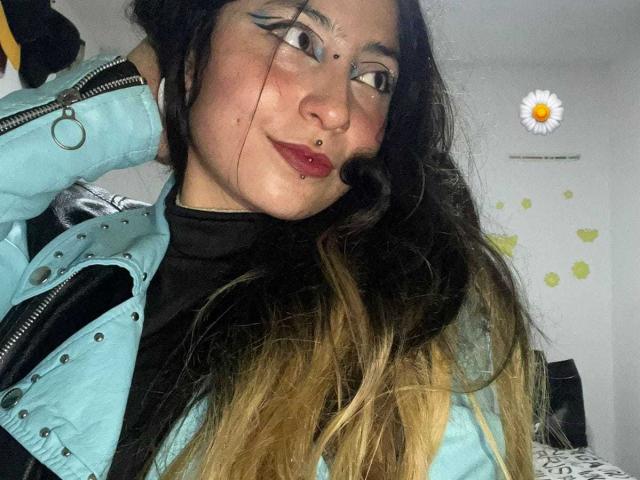 SakuraBlackRose, avec de longs cheveux noirs et blonds, portant une veste bleue, un t-shirt noir et du rouge à lèvres, a des piercings au nez et aux lèvres, et du crayon noir pour les yeux, et elle sourit légèrement..
