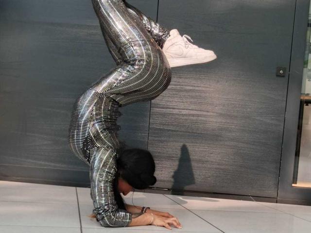 SakuraBlackRose staat op kop, met glanzende broek met een uitgesneden achterkant, witte sneakers en een zwarte pet, en doet een handstand tegen een donkergrijze muur..
