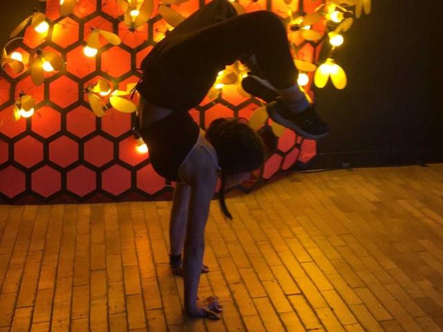 SakuraBlackRose sta eseguendo un'incredibile posizione di equilibrio a testa in giù, con i glutei rivolti verso l'alto, indossando pantaloncini neri e una canotta, con la testa tra le gambe su un pavimento di legno..