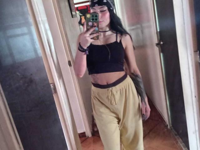 SakuraBlackRose with long black hair, black crop top, yellow pants, and tattoos, taking a mirror selfie, showing her flat stomach and small tits, wearing a black bandana.