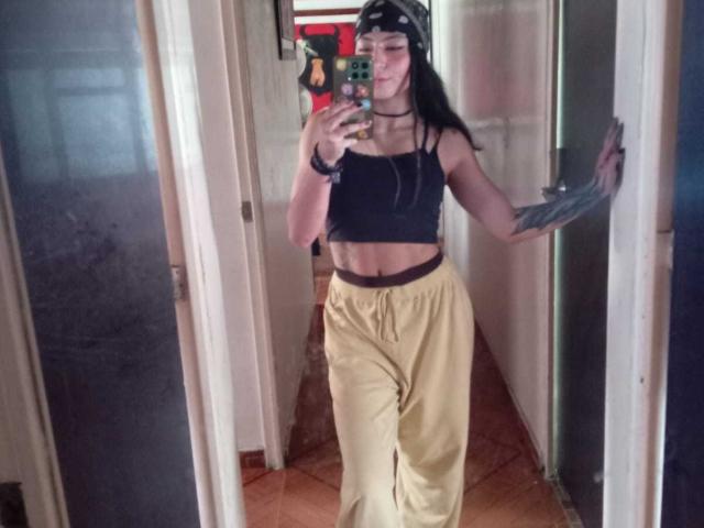 SakuraBlackRose con top nero, pantaloni gialli, tatuaggio sul braccio destro, bandana nera, che tiene un telefono, selfie allo specchio, magra, addome piatto, capelli lunghi neri, pavimento di legno, stile casual..