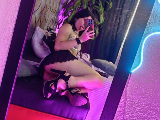 SakuraBlackRose, Spiegel-Selfie, kniend, schwarze High Heels, kurzes schwarzes Kleid, das ihren Hintern zeigt, Handy in der Hand, violettes Neonlicht, Pflanze im Hintergrund, schwarzer Armreif, pink-violette Beleuchtung..