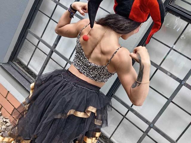 SakuraBlackRose, de costas para a câmera, exibe os músculos, vestindo uma blusa de estampa de leopardo, uma saia de tule preta com detalhes dourados, um chapéu de bobo da corte com cores vermelhas e pretas, e mostra uma tatuagem no braço direito..