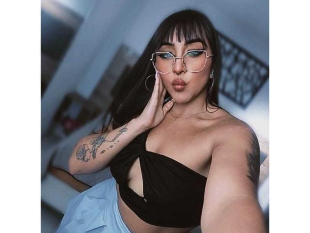 MissCharlottt with glasses, black wrap top showing her boobs, blue pants, tattoos on her arm, hand on cheek, pouting lips, dark hair, and hoop earrings, in a selfie.