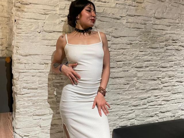 SakuraBlackRose trägt ein eng anliegendes weißes Kleid, das ihren Hintern und ihre kleinen Brüste zur Schau stellt, mit einem schwarzen Choker, Tattoos und Armbändern, und steht vor einer weißen Ziegelwand..