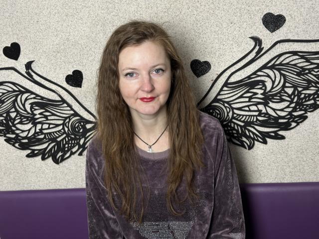 MeganPirs has wavy brown hair, red lipstick, and wears a purple velvet shirt with a necklace. Black wing tattoo is on the wall behind her.