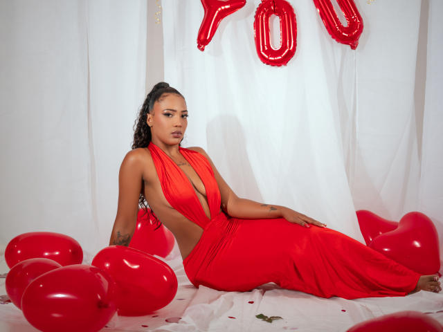 MorganaX with tan skin, black curly hair, wearing a red halter dress, lying on white cloth, surrounded by red balloons, showing her ass and sideboob.