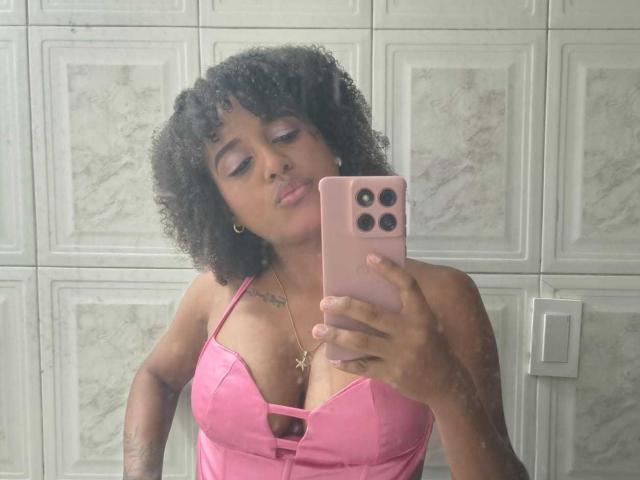 TentacionLatine, com cabelo preto encaracolado, vestindo uma blusa rosa de cetim que mostra seus seios, tirando uma selfie com um celular rosa, com uma expressão de lábios franzidos, tem uma tatuagem no peito..