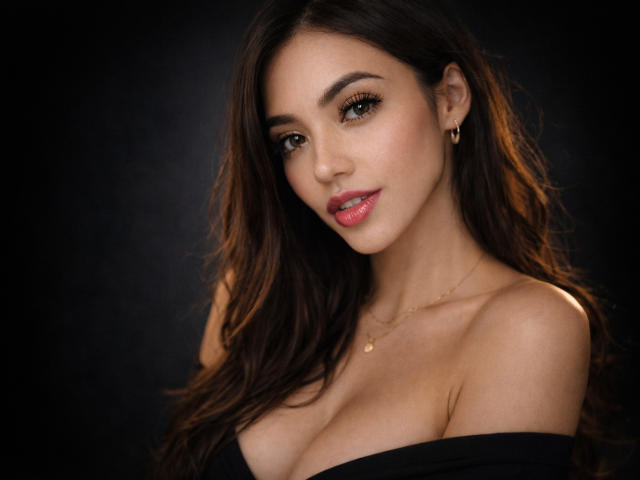SofieWhite with long brown hair, green eyes, and pink lipstick, wearing a black off-shoulder top, showing her tits, gold necklace, and earrings, against a dark background.