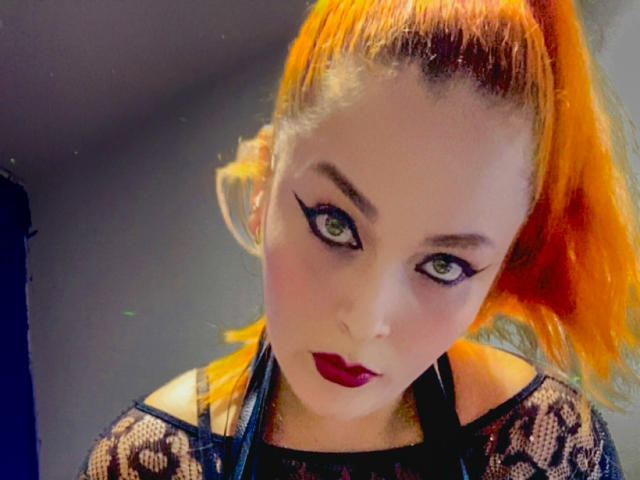 DominatrixMorrigan with bright orange ponytail, green eyes, black eyeliner, red lipstick, wearing black lace top, looking down with a slightly annoyed expression.