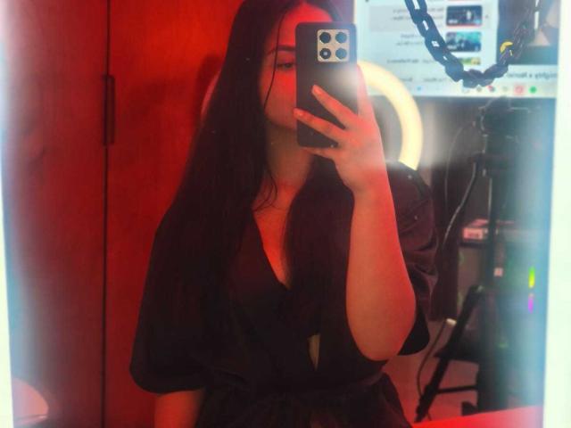 RouxiMori with long black hair, taking a mirror selfie, wearing a black dress, holding a phone, red lighting, visible ring light, and a chain in the background.