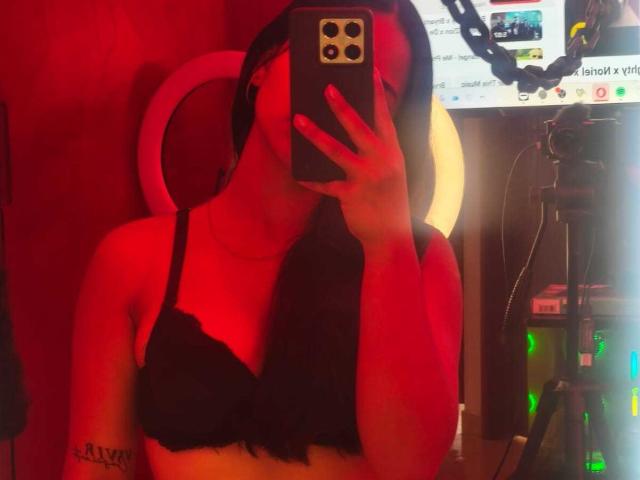 RouxiMori with red lighting, black lace bra, taking a mirror selfie with iPhone, visible tattoo on left arm, long black hair, holding phone over face, camera to right.
