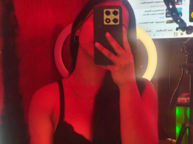 RouxiMori with long black hair, wearing a black lace bra, taking a selfie in red lighting, holding a phone with a gold camera, ring light behind her.
