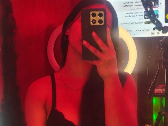 RouxiMori with long black hair, wearing a black lace top, holding a phone with a ring light in the background, taking a mirror selfie. Boobs visible. Red lighting.