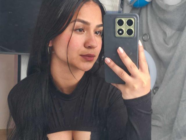 RouxiMori with long black hair, wearing a black long-sleeve top, holding a phone in front of her face, showing her tits, taking a selfie, with a slight smile.