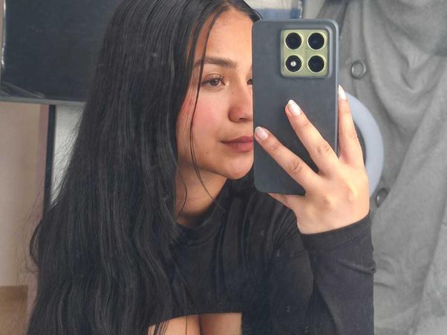 RouxiMori with long black hair, holding a green iPhone, taking a mirror selfie, wearing a black shirt, looking at the camera with a slight smile, visible nose and lips.