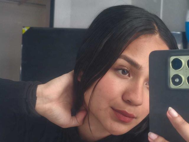 RouxiMori with black hair, holding a phone, showing off a cute face and slightly parted lips. Her hand's touching the back of her neck. She's wearing a black top.
