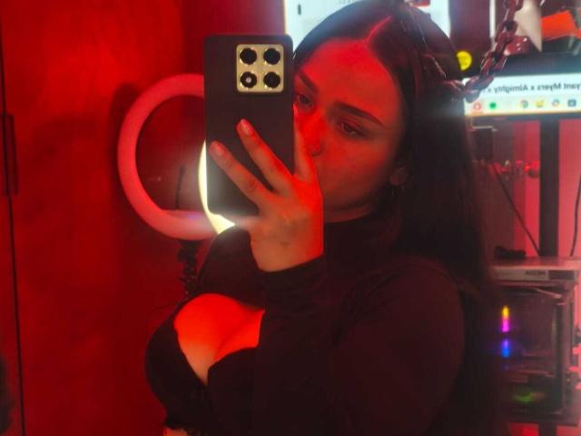 RouxiMori taking a mirror selfie with red lighting, holding a phone, wearing a black long-sleeve top, showcasing her large boobs, with long dark hair, looking at the camera.