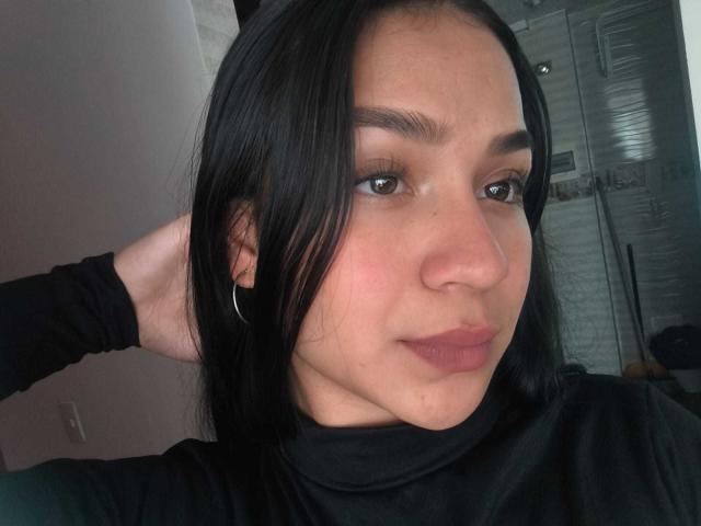 RouxiMori has long black hair, brown eyes, and a slight smile, wearing a black top with a hand behind her head, hoop earring, and pinkish-brown lipstick.