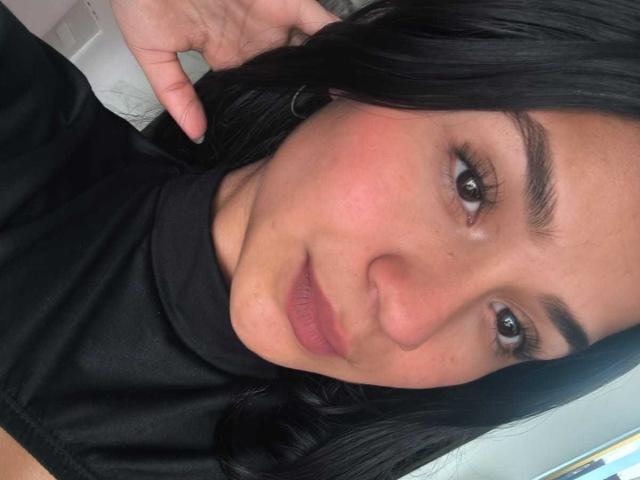 RouxiMori's face, showing a slight smile with pink lips, brown eyes, and black hair, wearing a black shirt, hand touching her head, looking at the camera.