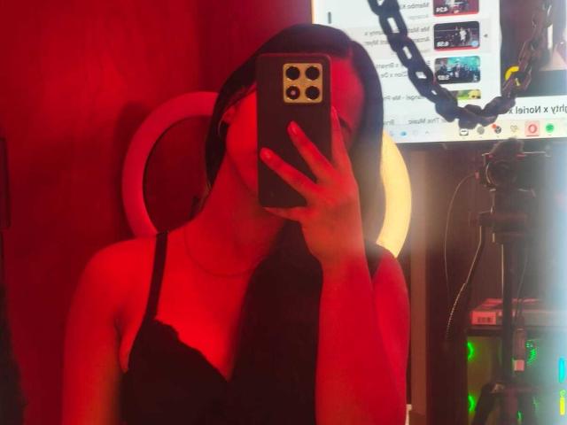 RouxiMori with red lighting, taking a selfie in a mirror, wearing a black lace top, covering her face with a phone, headphones on, in a dimly lit room.