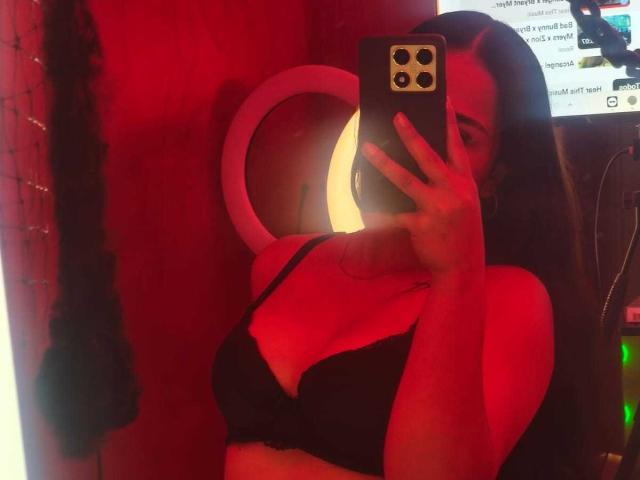 RouxiMori with red lighting, taking a mirror selfie, wearing a black bra, holding a phone, covering her face, long brown hair, visible cleavage, ring light in background.