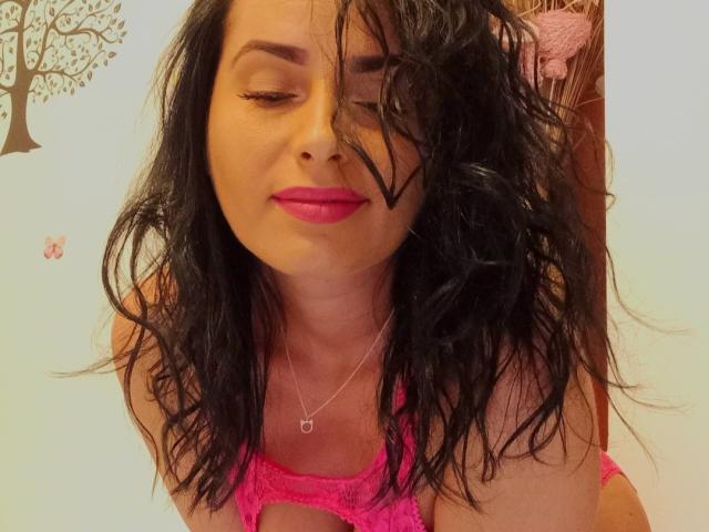 MisstyX with wavy black hair, pink lipstick, and a pink lace bra, leaning forward, eyes closed, wearing a silver heart necklace, smiling slightly. Boobs visible, nipples hidden.