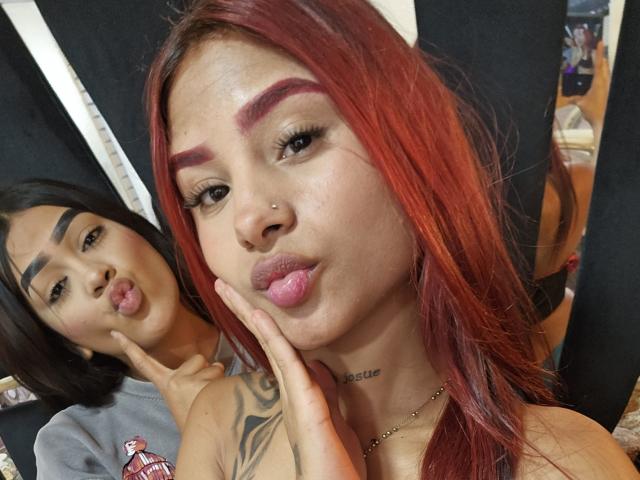 TatianaYesica with red hair and tattooed arm, pouting lips, and pierced nose, stands next to TatianaYesica with black hair, sticking out tongue, making "shush" gesture.