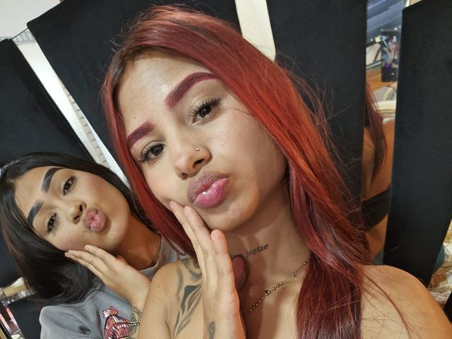 TatianaYesica with red hair, pouty pink lips, and tattooed neck, holding her chin. TatianaYesica with black hair, pouting, in background. Boobs visible under TatianaYesica's top.