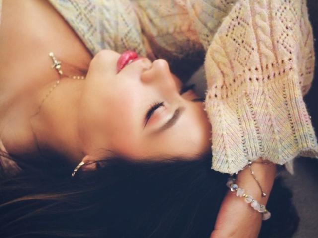 AshleyyPort, con gli occhi chiusi, indossa un maglione di lana, orecchini dorati e un braccialetto di perline, è sdraiata con una mano sulla testa e ha il rossetto rosa..