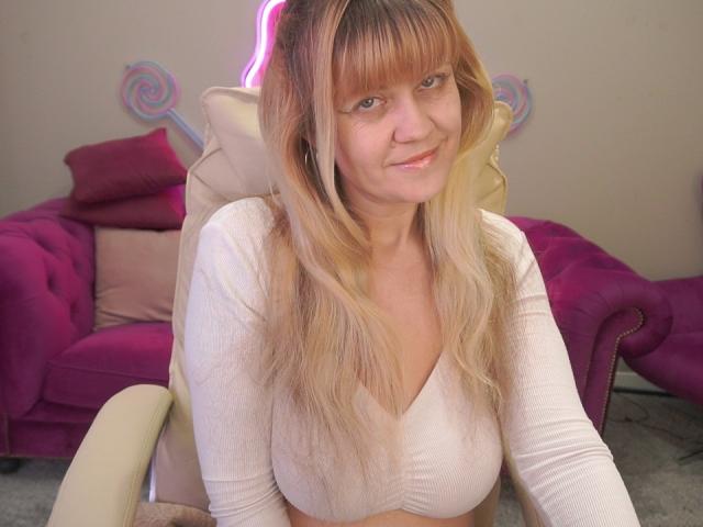 BellaLaPetale, a middle-aged woman with light skin and blonde hair with bangs, sits in a beige chair. She has a subtle smile and wears a white, long-sleeve crop top that reveals her midriff. The background includes a pink plush sofa, a purple cushion, and decorative swirls on the wall. Her hair is wavy and she has a natural, slightly tired look. The image has a soft, casual vibe.