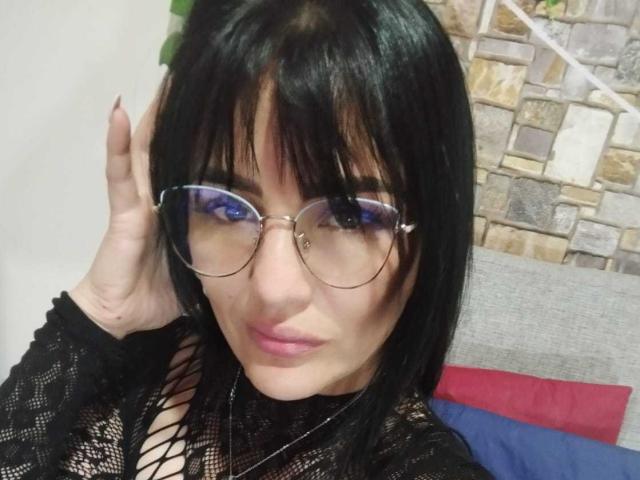 CrazyRousesex with black hair, glasses, and pink lipstick, wearing a black lace top with fishnet, touching her head, showing her tits through the lace.