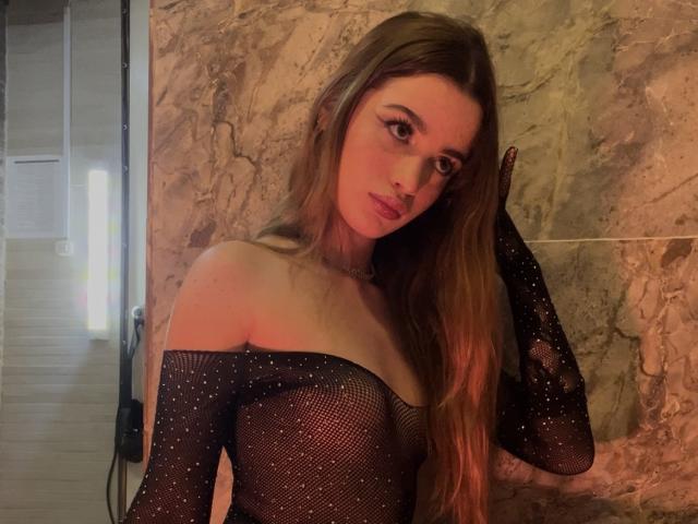AprillAhego with long brown hair, wearing a sparkly black mesh off-shoulder top, showing her small tits, and a subtle pout, against a marble wall.