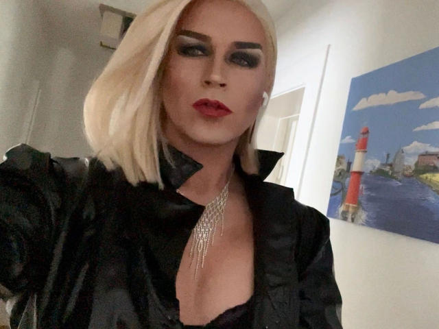 CassyTV, con un corte de pelo rubio platino, maquillaje negro, lápiz labial rojo, una camisa de satén negro, escote pronunciado, un collar de plata y un collarín negro, mostrando sus senos..
