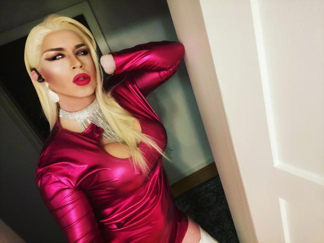 CassyTV, aantrekkelijk in een glanzend roze metallic top met een decolleté, lang blond haar, rode lippenstift en een diamanten ketting, staat bij een deur, met haar hand op haar hoofd..