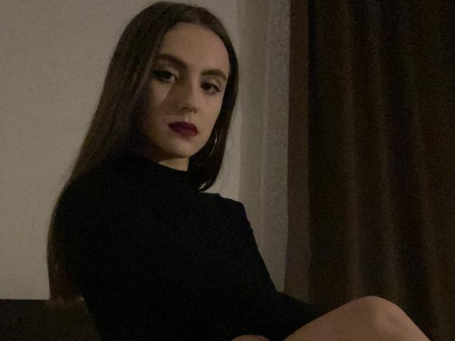 MaddymidNight with long brown hair, dark lipstick, and black turtleneck, sitting with legs crossed, looking slightly to the side, in dim lighting. Boobs not visible, pussy not visible.