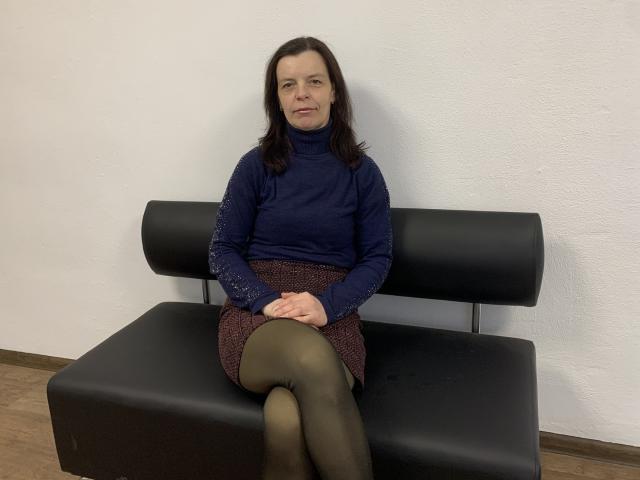 SofiaChaika zit op een zwarte leren bank met gekruiste benen, ze draagt een blauwe coltrui, een bruin geruite rok en zwarte panty's, en ze kijkt er iets van verveeld uitzien met haar gladde bruine haar..