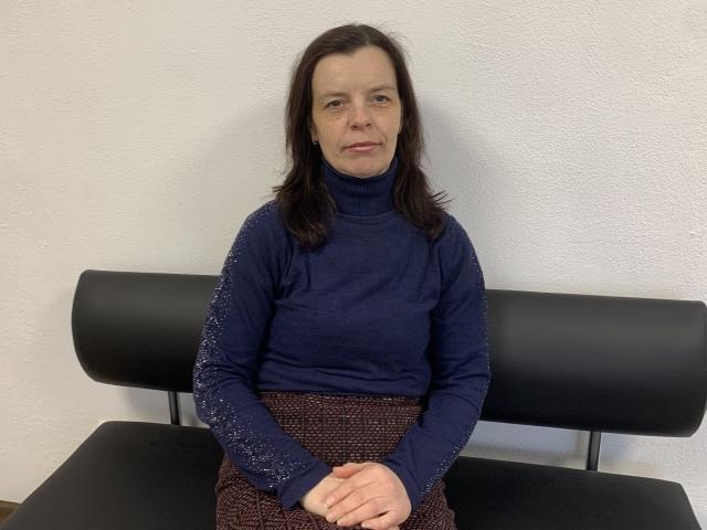 Sofia Chaika, con cabello castaño de longitud media, vestida con un cuello alto azul y mangas brillantes, y una falda marrón estampada, sentada en un banco de cuero negro, con las manos en el regazo..