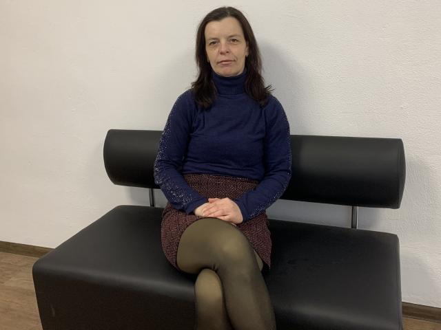Sofia Chaika, com seios médios, vestindo uma gola alta azul, uma saia escura e collant verdes, sentada em um banco preto, com as pernas cruzadas, em um quarto simples..