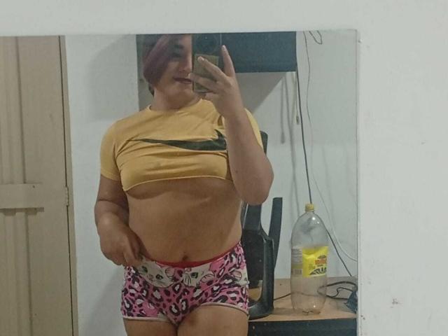 MiaSims mit kurzen, lila Haaren, einem gelben Crop-Top, das ihre Brüste zeigt, pinken Shorts mit Leopardenmuster, stehend vor einem Spiegel, ein Handy haltend, lächelnd, mit sichtbarem Bauchnabel..