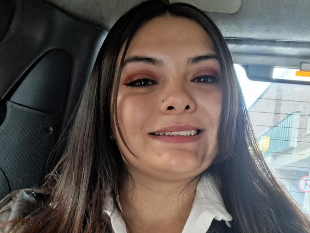 KatherinLatin a les cheveux bruns, un fard à paupières rouge, un eye-liner noir, un piercing au nez, et elle sourit en montrant ses dents. Elle porte une chemise blanche et se trouve dans une voiture..