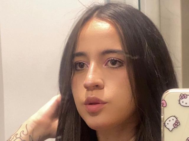 Close-up of IrissMontiel with long, straight black hair, light brown skin, and pinkish-brown lips. She has small, dark eyebrows, slightly arched, and brown eyes. Her right hand is touching her hair. She's holding a phone with a Hello Kitty sticker case in her left hand. A small tattoo is visible on her right wrist. The image has a slightly glossy texture. IrissMontiel's expression is neutral.