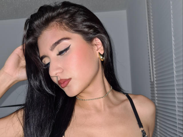 NaylaGrey with long black hair, wearing gold earrings and a black bra, has bold eyeliner, pink blush, and red lipstick, looking down with a slight smile, hand in hair.