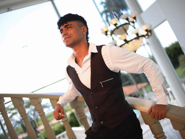 Jacobb69 leans on a wooden railing, facing left with a slight smile. He has curly black hair and a medium-brown skin tone. He wears a white shirt with a black vest that has a pocket. The background shows a chandelier and glass windows. The light is bright, highlighting his confident posture. The composition focuses on Jacobb69's relaxed, yet poised, stance.