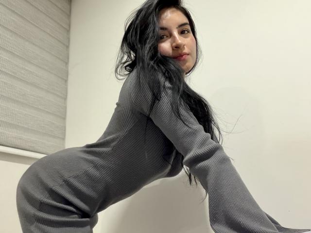 EmilyJensenn with long black hair, wearing a gray ribbed dress, leaning forward, showing her ass, looking at the camera with a slight smile.