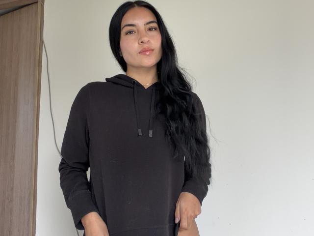 EmilyJensenn with long black hair, wearing a black hoodie, standing with hands in pockets, smiling slightly, medium brown skin, cute, no makeup, natural look, wooden door on left.